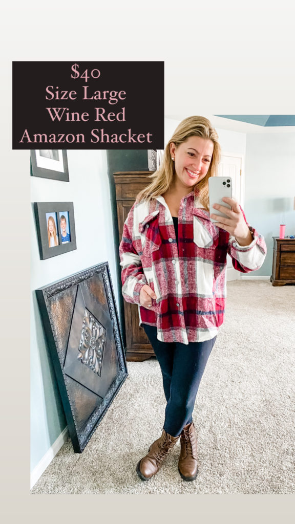 A woman looks into her phone while smiling in the mirror. Her legs are crossed and her hand is in the pocket of her shirt. She is wearing a wine colored flannel shirt jacket 'shacket' with black leggings and brown combat boots. The text on the photo says $40 size large wine red amazon shacket. 