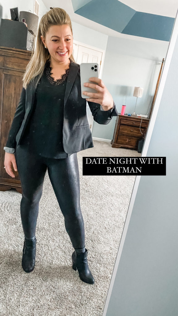 A picture of a woman wearing an all black outfit while holding her phone and smiling in the mirror. She is wearing a black blazer, a black lace tank, faux leather leggings, and black booties with a heel. The caption on the photo says Date Night with Batman.