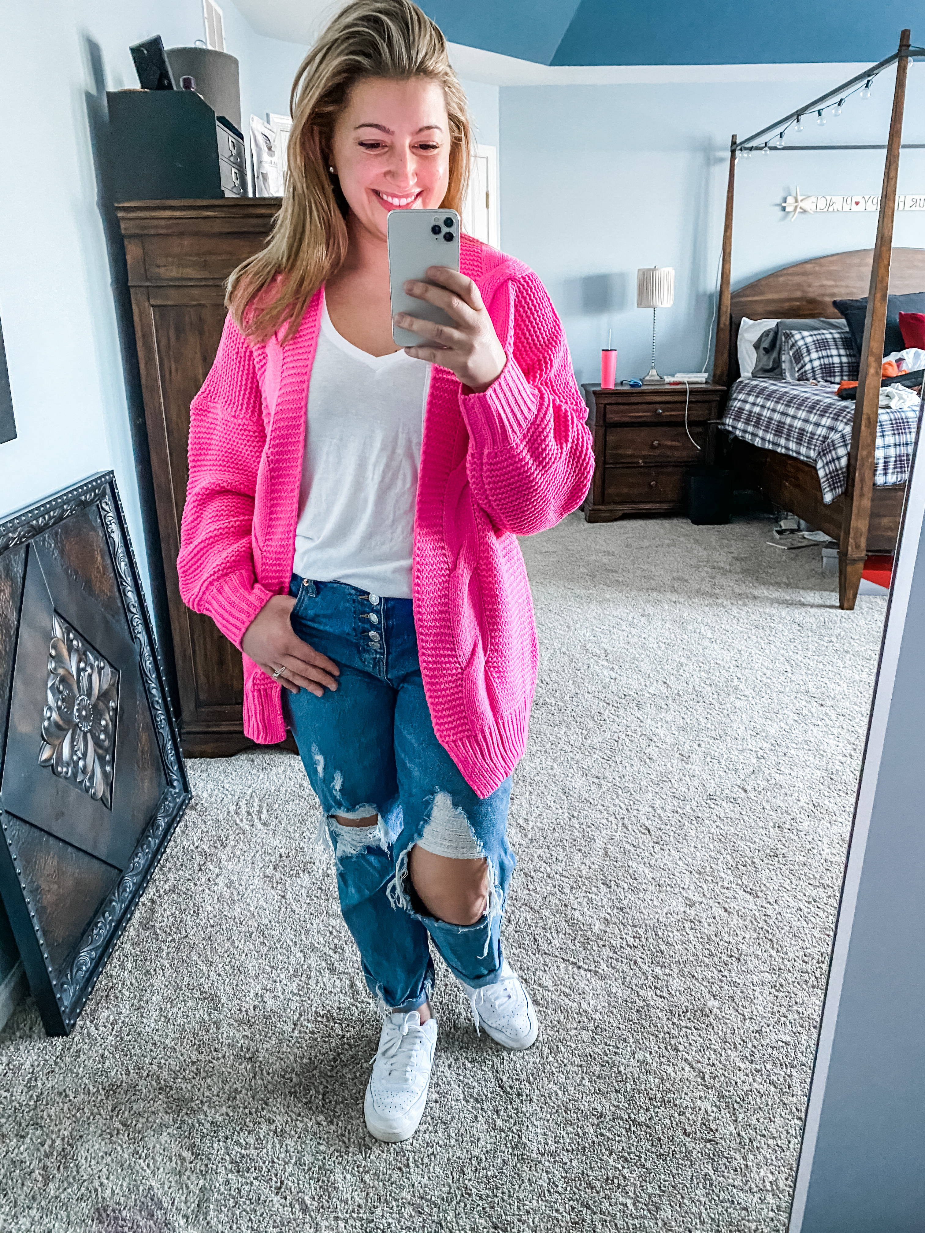A young woman with blonde hair smiles in the mirror as she holds her camera. She is wearing a hot pink long cardigan, white tank top, distressed jeans and white sneakers. 