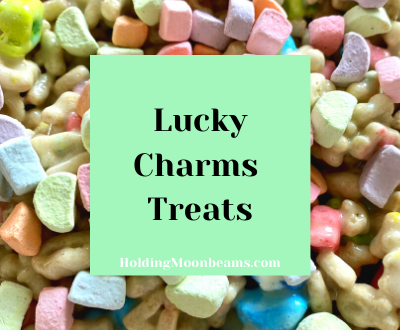 It's a large picture of Lucky Charms cereal pieces - there are purple moons, rainbows, red stars, and others that make up the background image. In the center of the photo, there is a green square that states 'Lucky Charms Treats' in bold black letters. Within the green box, there is a line at the bottom that says "HoldingMoonbeams.com" This image is used as an blog graphic to visually show what the rest of the post is about which is a Lucky Charms Treats recipe and directions.