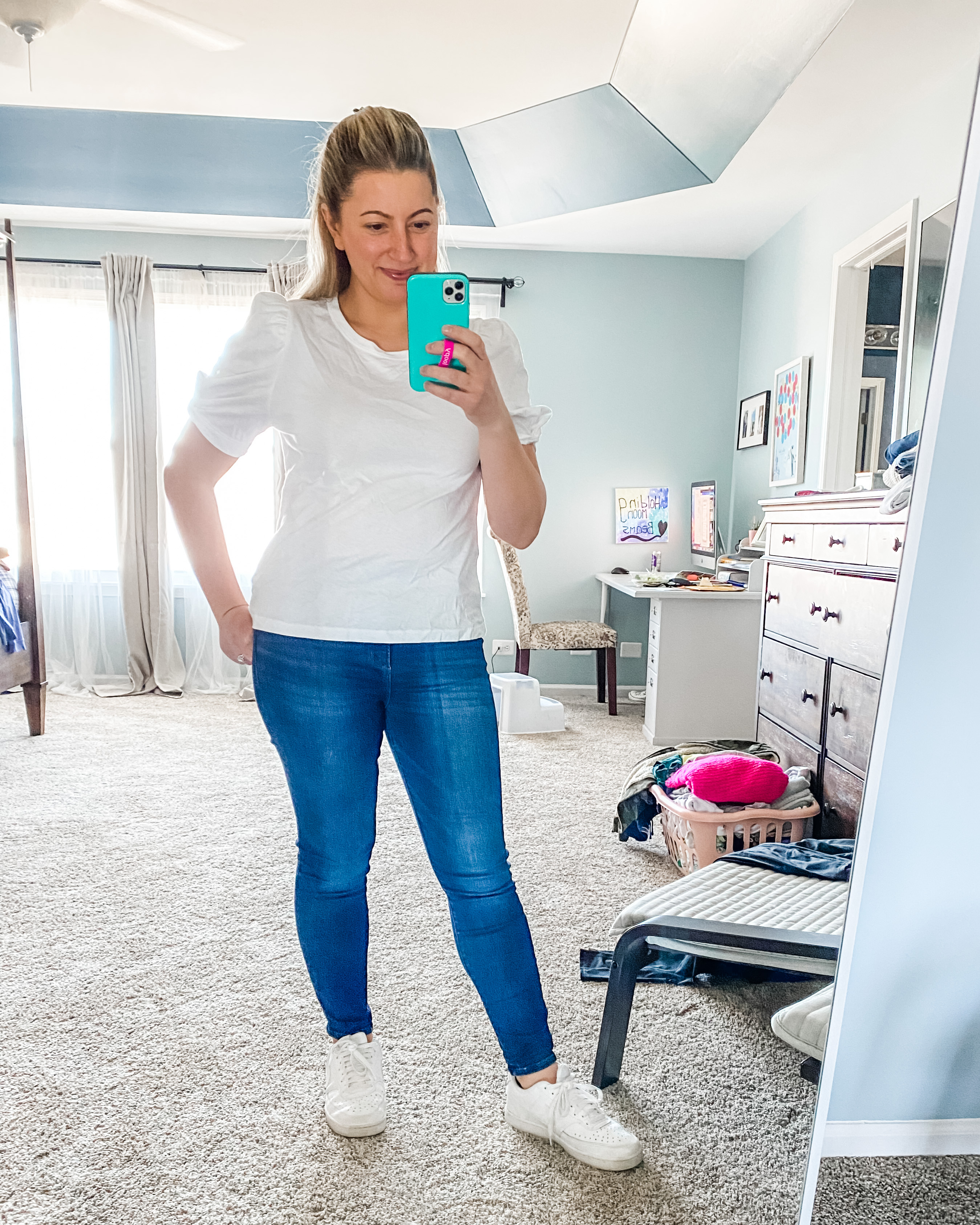 A young female in her 30s is standing in her bedroom with a messy background of furniture and clothes. She has blonde hair pulled back, is wearing a puff sleeved T-shirt that is white. Medium wash blue denim jeans, and Nike white sneakers. She is holding her phone in one hand and the other hand is on her hip. She is smiling and looking into the phone. It appears that she’s taking the picture of her self in the mirror. If you click on the picture, it takes you to a link where you can shop the items in the photo.