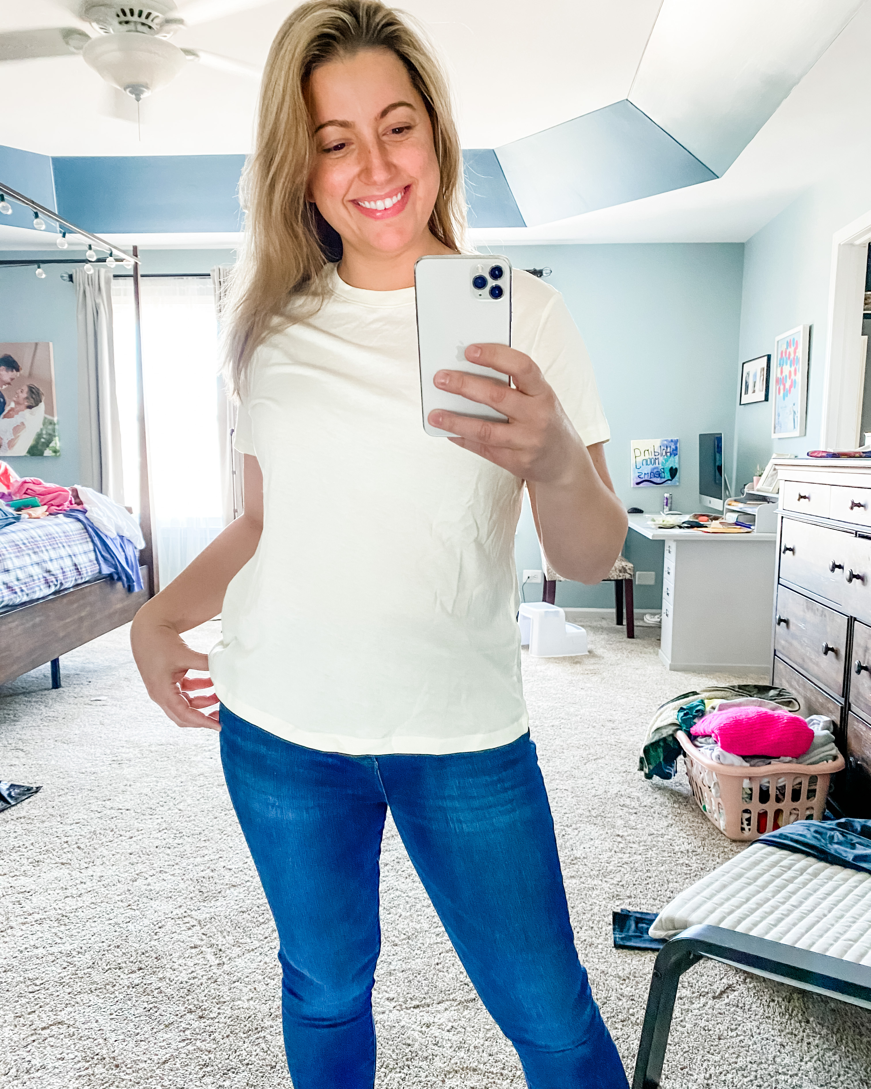 A young female in her 30s is standing in her bedroom with a messy background of furniture and clothes. She is holding her iPhone in one hand and has one hand  on her hip. She has a medium length blonde hair and is smiling while she’s looking into the phone. She is wearing a short-sleeved yellow crewneck T-shirt from target with medium wash blue denim jeans from Abercrombie. If you click on the picture, it takes you to a link where you can shop the items in the photo.