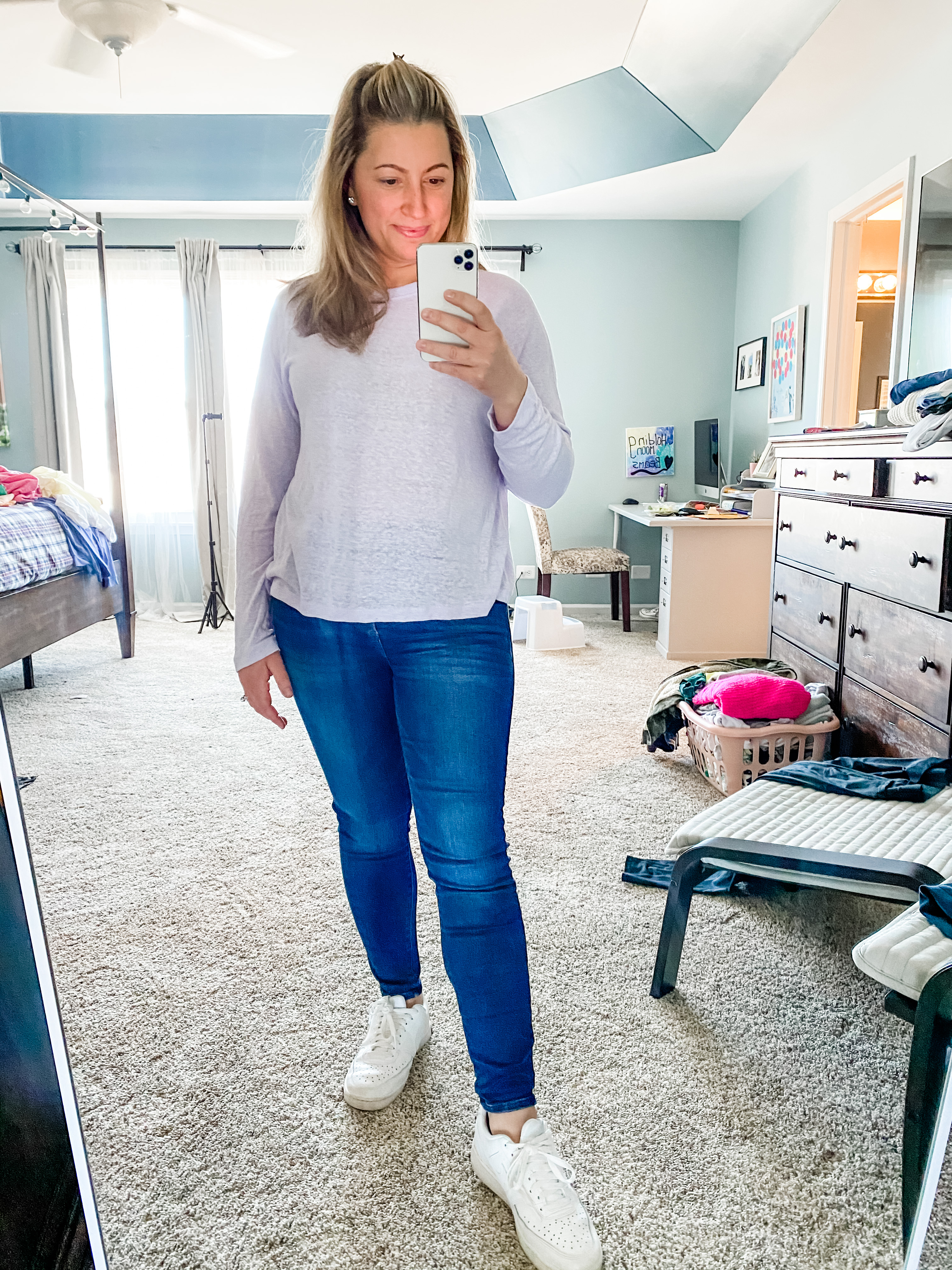 A young female in her 30s is standing in her bedroom with a messy background of furniture and clothes. She is standing full length and a long mirror. She has a pursed smile as she looks into the phone. Her other hand is down by her hip. She is wearing a long sleeve lilac purple shirt that is sheer. Her hair is half up. She’s wearing blue denim jeans with white sneakers. The reflection of the phone glows on her face. She is standingShe is holding her iPhone in one hand and has one hand  on her hip. She has a medium length blonde hair and is smiling while she’s looking into the phone. She is wearing a short-sleeved yellow crewneck T-shirt from target with medium wash blue denim jeans from Abercrombie.If you click on the picture, it takes you to a link where you can shop the items in the photo.