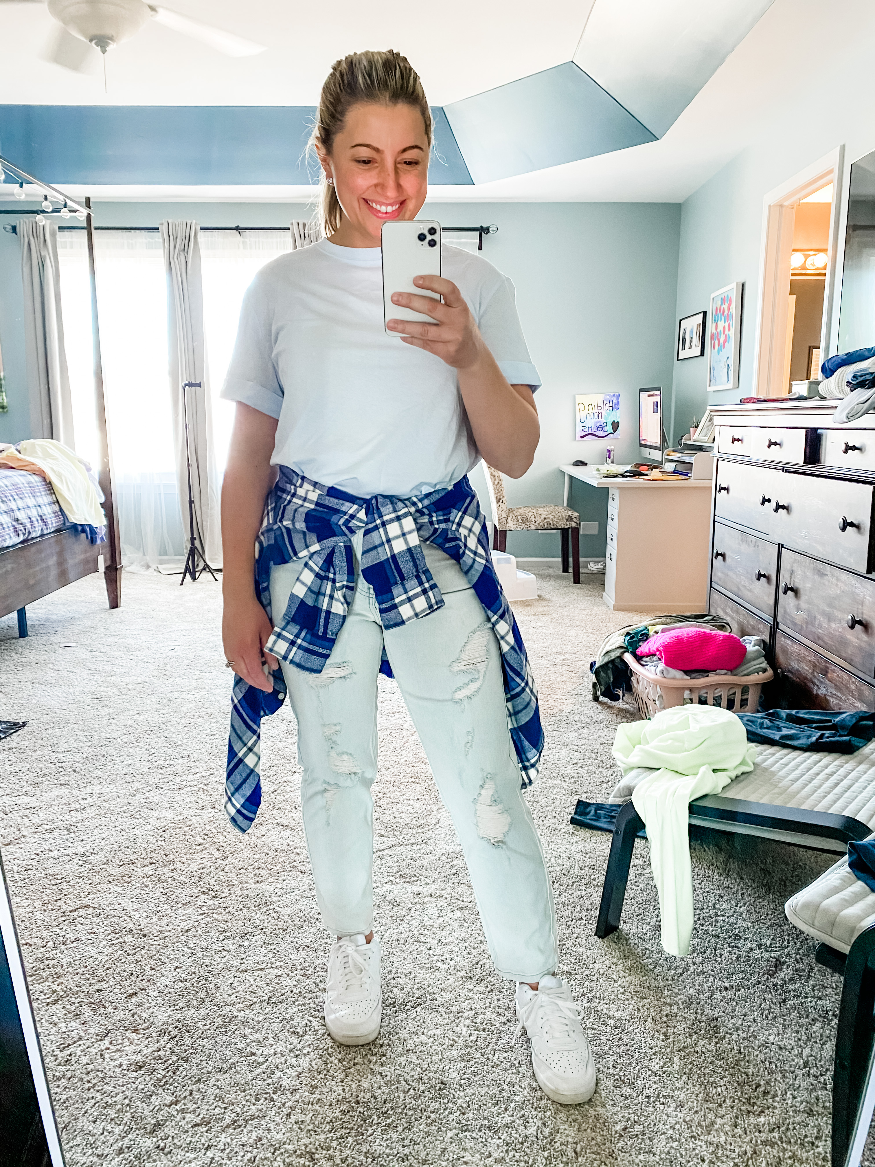 A young female in her 30s is standing in her bedroom with a messy background of furniture and clothes. She is wearing a light blue short sleeve shirt. The sleeves have cuffs. She has a blue and white checkered plaid flannel wrapped around her waist. She is wearing a light blue distressed mom jeans from target. And her favorite white sneakers. One hand is holding the phone in the other is down by her side. She is smiling at the phone. PhoneIf you click on the picture, it takes you to a link where you can shop the items in the photo.
