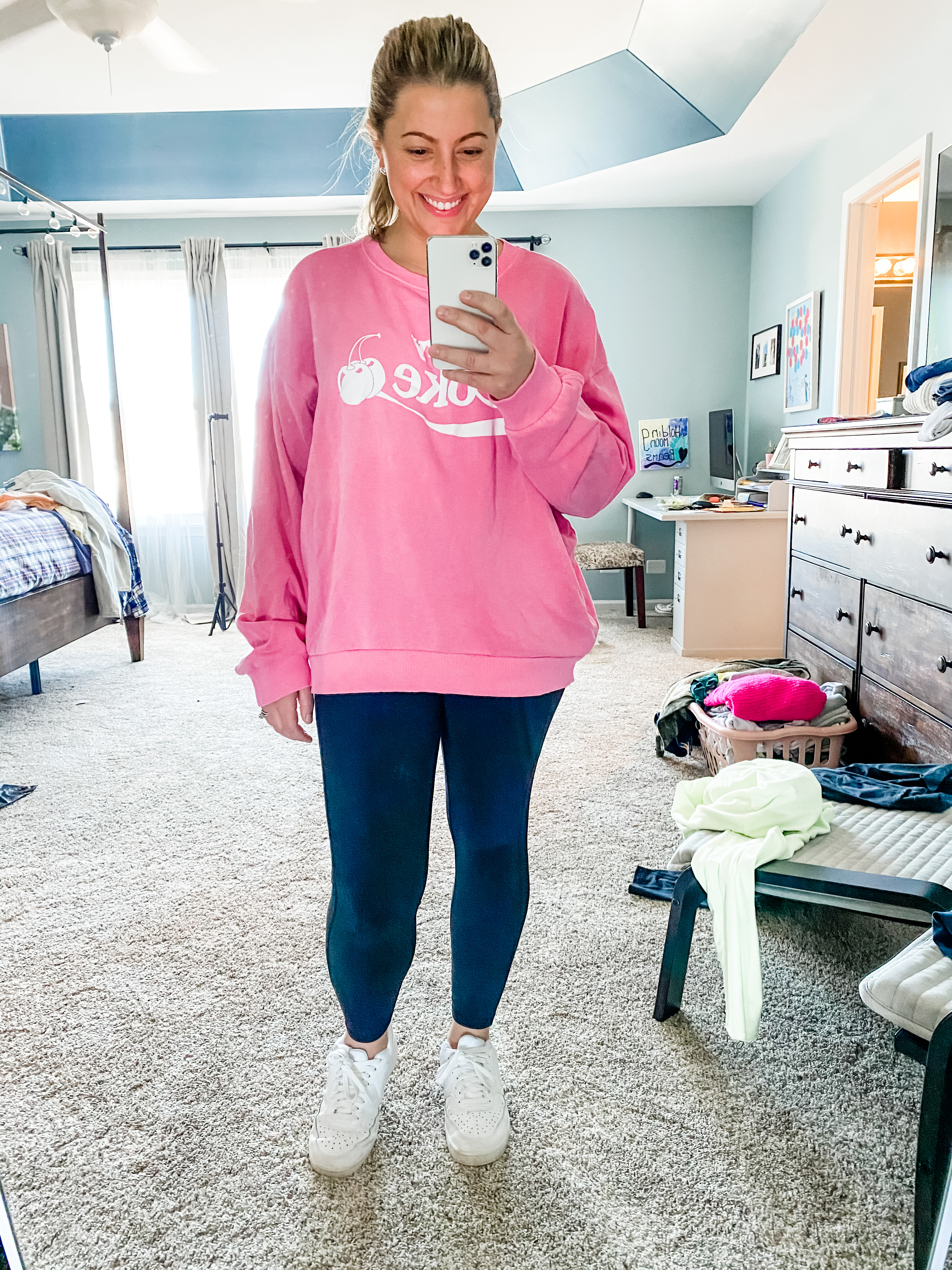 A young female in her 30s is standing in her bedroom with a messy background of furniture and clothes. Her hair is in a ponytail and she is wearing an oversized pink sweatshirt that says cherry Coke with two cherries in white writing. She is wearing black stretchy leggings and white sneakers. She is smiling into the phone as if she’s taking this picture in a mirror.If you click on the picture, it takes you to a link where you can shop the items in the photo.